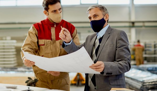 Mid adult businessman and manual worker analyzing plans while working at carpentry workshop during coronavirus pandemic.